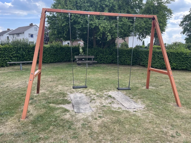 Ein Spielplatz mit einem Holzschaukelrahmen und zwei Schaukeln auf einer Wiese. Hintergrund: Büsche.