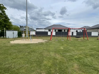 Spielplatz Specker Straße Ein Rasenplatz mit Klettergerüst und roter Schaukel, dahinter weiße Gebäude.