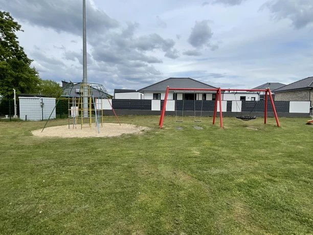 Spielplatz Specker Straße Ein Rasenplatz mit Klettergerüst und roter Schaukel, dahinter weiße Gebäude.