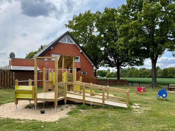 Auf dem Bild ist ein freundlicher Spielplatz zu sehen. Eine Holzrampe führt zu mehreren Spielgeräten. Im Hintergrund steht ein rotes Backsteinhaus unter hohen Bäumen.