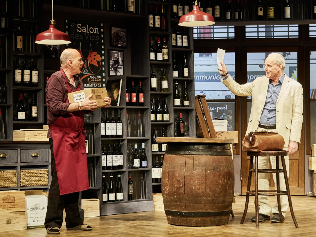 Weinprobe für Anfänger, Neues Theater Zwei ältere Männer stehen in einem Weingeschäft. Einer hält ein Glas Wein, der andere eine Karte.