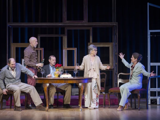 Und wenn wir alle zusammenziehen, Neues Theater Das Bild zeigt eine Theaterszene mit fünf Personen, elegant gekleidet, die lebhaft an einem Tisch diskutieren.