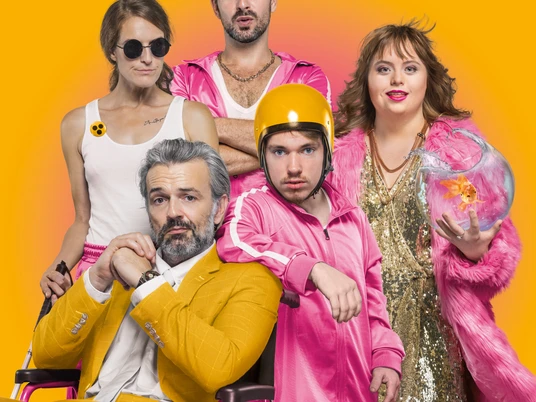 Die Goldfische, Neues Theater Eine Gruppe von fünf Menschen, gekleidet in knalligen Farben, posiert lebhaft vor einem leuchtend gelben Hintergrund. Eine Person im Rollstuhl trägt einen gelben Anzug, während die anderen in auffälligen rosa Outfits gekleidet sind. Einer trägt einen goldenen Helm, eine Person eine goldene Glitzerjacke und hält einen Goldfisch in einem Glas.