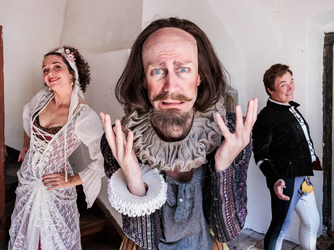 Es war die Lerche Drei fröhlich gekleidete Schauspieler posieren in historischen Kostümen vor hellen Wänden.