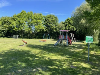 Spielplatz Zur Alten Schule Auf dem Bild ist ein Spielplatz mit einem bunt gestalteten Klettergerüst, Schaukeln und einem Wipptier zu sehen. Im Hintergrund umsäumen Bäume eine offene Grünfläche, die an einem sonnigen Tag das Areal umrahmen.