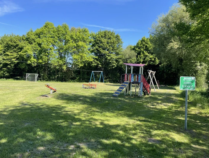 Spielplatz Zur Alten Schule Auf dem Bild ist ein Spielplatz mit einem bunt gestalteten Klettergerüst, Schaukeln und einem Wipptier zu sehen. Im Hintergrund umsäumen Bäume eine offene Grünfläche, die an einem sonnigen Tag das Areal umrahmen.