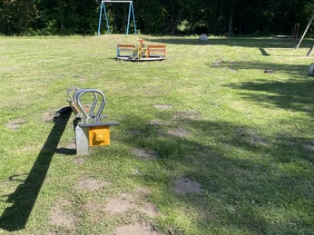 Grüner Spielplatz mit Wiese, Schaukel, Wippe und Klettergerüst; umgeben von Bäumen.