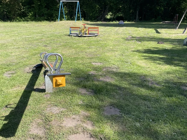 Spielplatz Zur Alten Schule Grüner Spielplatz mit Wiese, Schaukel, Wippe und Klettergerüst; umgeben von Bäumen.