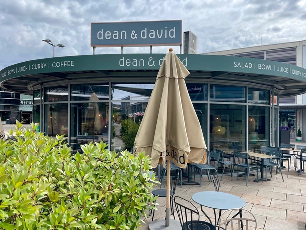Man sieht den runden Glas-Pavillon der dean & david Filiale in den Designer Outlets Wolfsburg. Davor ist der Außensitzbereich mit Tischen und Stühlen.