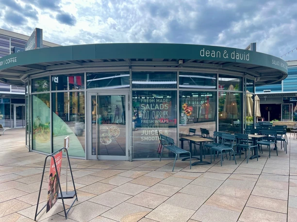 Man sieht den runden Glas-Pavillon der dean & david Filiale in den Designer Outlets Wolfsburg. Mittig ist die Eingangstür und rechts der Außensitzbereich mit Tischen und Stühlen.