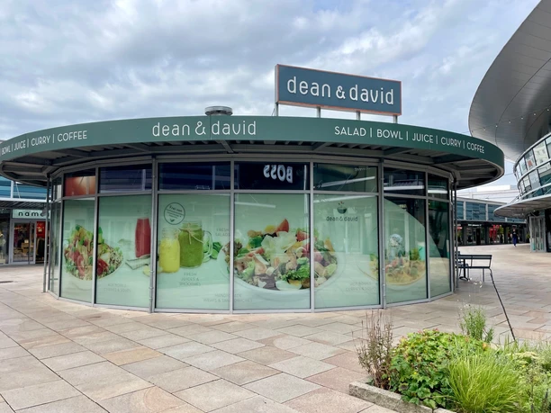 Man sieht die Rückseite des runden Glas-Pavillon der dean & david Filiale in den Designer Outlets Wolfsburg. Die Glasfassade ist mit einer großflächigen Werbung beklebt, die die angebotenen Speisen präsentiert.