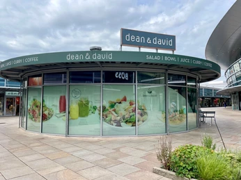 dean and david 4.jpg Man sieht die Rückseite des runden Glas-Pavillon der dean & david Filiale in den Designer Outlets Wolfsburg. Die Glasfassade ist mit einer großflächigen Werbung beklebt, die die angebotenen Speisen präsentiert.