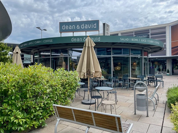 Man sieht den runden Glas-Pavillon der dean & david Filiale in den Designer Outlets Wolfsburg. Davor ist der Außensitzbereich mit Tischen und Stühlen.