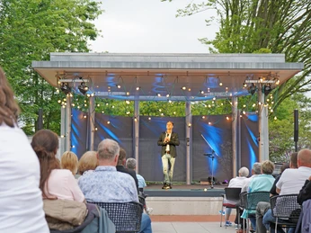 Thieß Neubert.png Mann steht mit Mikrofon auf beleuchteter Bühne und das Publikum schaut auf ihn