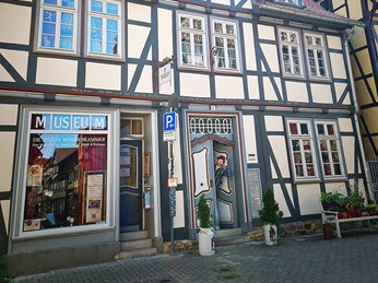 Haus Sommer25.jpg Fachwerkhaus mit weiß-blauen Fassaden, Museumsschild und Pflanzen am Eingang, blauer Himmel.