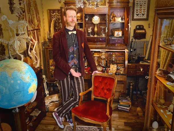 Ein Mann in einem stilvollen, vintage-inspirierten Raum zeigt ein hohes, rotes Polstersessel. Breite Holzmöbel, ein Globus und skurrile Sammlerstücke zieren den Hintergrund. Sein Outfit mit Fliege und gestreiften Hosen verstärkt den alten Charme des Zimmers.