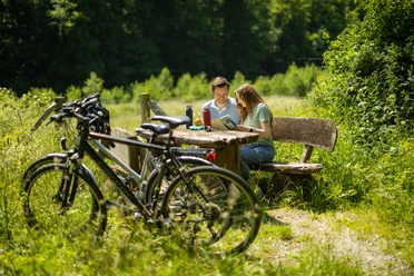 Ein Paar sitzt an einem Holztisch im Grünen, umgeben von Fahrrädern und dichter Vegetation.