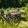 Paderborner-Land_Radfahren_Altenau_009 ©Kreis Paderborn, Sabrinity.jpg Ein Paar sitzt an einem Holztisch im Grünen, umgeben von Fahrrädern und dichter Vegetation.