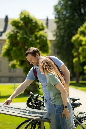 Paderborner-Land_Radfahren_Dalheim_013 ©Kreis Paderborn, Sabrinity.jpg Ein Paar betrachtet eine Infotafel mit Fahrrad im Grünen, umgeben von Bäumen und einem Gebäude.