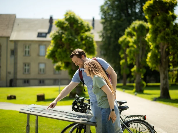 Paderborner-Land_Radfahren_Dalheim_013 ©Kreis Paderborn, Sabrinity.jpg Ein Paar betrachtet eine Infotafel mit Fahrrad im Grünen, umgeben von Bäumen und einem Gebäude.
