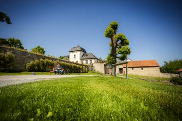 Paderborner-Land_Radfahren_Dalheim_020 ©Kreis Paderborn, Sabrinity.jpg Radfahrer genießen eine Pause auf einer Bank vor historischem Kloster Dalheim bei strahlendem Sonnenschein.