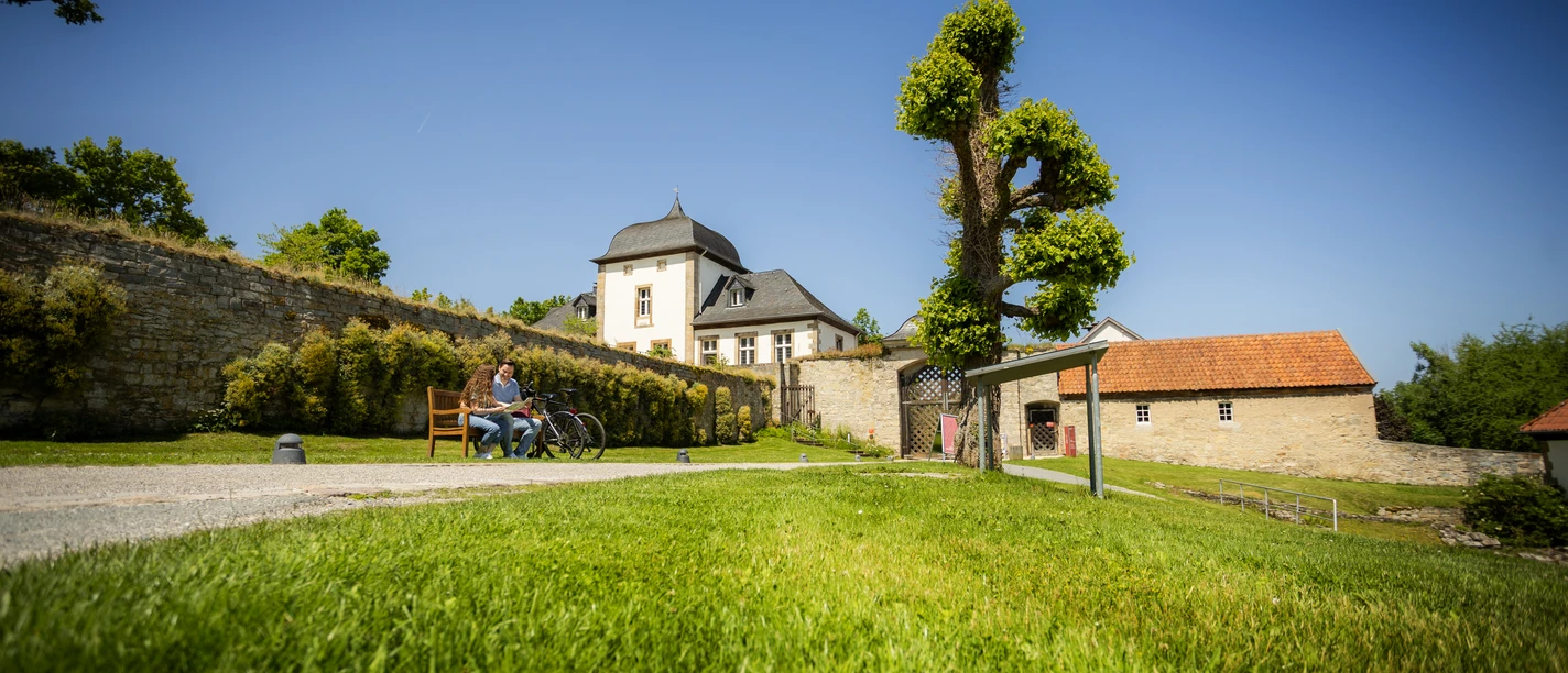 Paderborner-Land_Radfahren_Dalheim_020 ©Kreis Paderborn, Sabrinity.jpg Radfahrer genießen eine Pause auf einer Bank vor historischem Kloster Dalheim bei strahlendem Sonnenschein.
