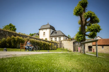 Paderborner-Land_Radfahren_Dalheim_021 ©Kreis Paderborn, Sabrinity.jpg Radfahrer auf einer Bank in ländlicher Kulisse vor historischem Gebäude mit verziertem Dach.