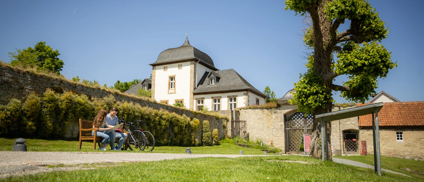 Paderborner-Land_Radfahren_Dalheim_021 ©Kreis Paderborn, Sabrinity.jpg Radfahrer auf einer Bank in ländlicher Kulisse vor historischem Gebäude mit verziertem Dach.