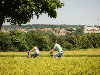 Zwei Radfahrer fahren durch eine grüne Landschaft mit Blick auf die Wewelsburg im Hintergrund.