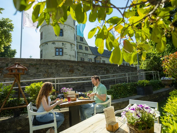 Zwei Menschen genießen eine Mahlzeit im Freien vor der historischen Wewelsburg bei Sonnenschein.