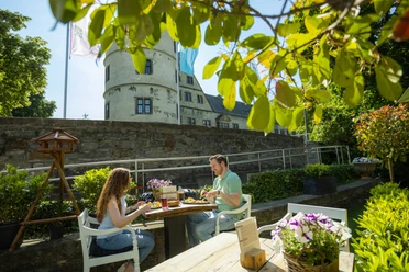 Paderborner-Land_Radfahren_Wewelsburg_Gastronomie_002 ©Kreis Paderborn, Sabrinity.jpg Zwei Menschen genießen eine Mahlzeit im Freien vor der historischen Wewelsburg bei Sonnenschein.