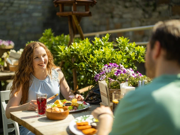Frau und Mann genießen ein Mittagessen im Freien an einem Holztisch mit grünem Hintergrund.