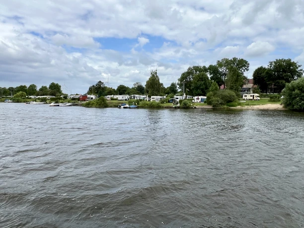 Campingplatz am Fluss mit Wohnwagen, Bäumen und einem Strand, unter einem bewölkten Himmel.
