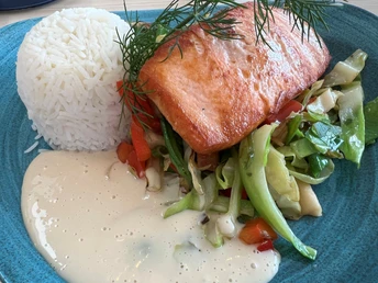 Lachsfilet auf frischem Gemüse mit Reis, dekoriert mit Dill, angerichtet auf einem blauen Teller.