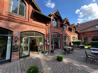 Exterieur eines Gästehauses in rotem Backsteingebäude mit großer Terrasse und Tischen im Freien.