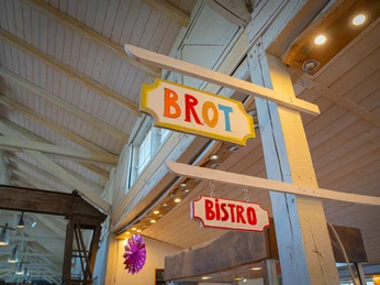 Firsches Brot und leckere Speisen Im Bioladen Moorrübe hängt eine bunte Retro-Schildwerbung für Brot und Bistro an einer weißen Holzkonstruktion. Die warme Beleuchtung und das Holzgebälk schaffen eine einladende Atmosphäre.