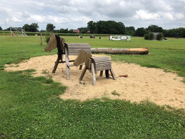 Two wooden play horses stand on a sandy area in the countryside under a slightly cloudy sky.