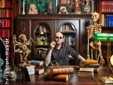 Ein Mann mit Sonnenbrille sitzt an einem Holztisch in einer Bibliothek voller Bücher und Kuriositäten.