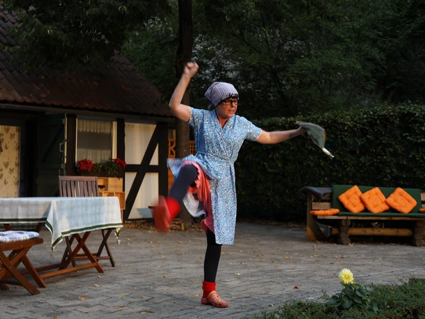 IMG_6584_17A2F266.JPG Eine Frau in einem bunten Kleid tanzt lebhaft auf einer Terrasse mit Gartenmöbeln im Freien.
