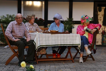 IMG_6606_9A979996.JPG Vier Personen in bunten Kostümen sitzen an einem Tisch im Freien, umgeben von Pflanzen.