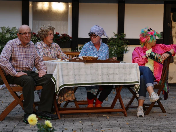 IMG_6606_9A979996.JPG Vier Personen in bunten Kostümen sitzen an einem Tisch im Freien, umgeben von Pflanzen.