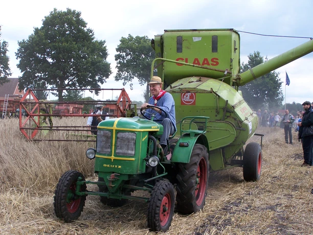 Kornmähen Aperberg Ein älterer Mann fährt einen kleinen grünen Deutz-Traktor, der einen großen, grünen CLAAS-Mähdrescher durch ein Getreidefeld zieht. Im Hintergrund stehen Zuschauer und weitere landwirtschaftliche Maschinen