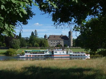 Das Passagierschiff Flotte Weser fährt auf einem Fluss inmitten einer grünen Landschaft vorbei an einem historischen Gebäude mit Türmen.The passenger ship Flotte Weser sails on a river in the middle of a green landscape past a historic building with towers.Passagerskibet Flotte Weser sejler langs en flod midt i et grønt landskab forbi en historisk bygning med tårne.Het passagiersschip Flotte Weser vaart over een rivier midden in een groen landschap langs een historisch gebouw met torens.