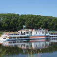 Das Ausflugsschiff Flotte Weser fährt auf einem Fluss, gesäumt von grünen Bäumen.The excursion boat Flotte Weser sails along a river lined with green trees.Udflugtsbåden Flotte Weser sejler langs en flod, der er omkranset af grønne træer.De excursieboot Flotte Weser vaart langs een rivier met groene bomen.