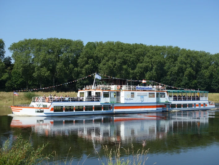 Das Ausflugsschiff Flotte Weser fährt auf einem Fluss, gesäumt von grünen Bäumen.The excursion boat Flotte Weser sails along a river lined with green trees.Udflugtsbåden Flotte Weser sejler langs en flod, der er omkranset af grønne træer.De excursieboot Flotte Weser vaart langs een rivier met groene bomen.