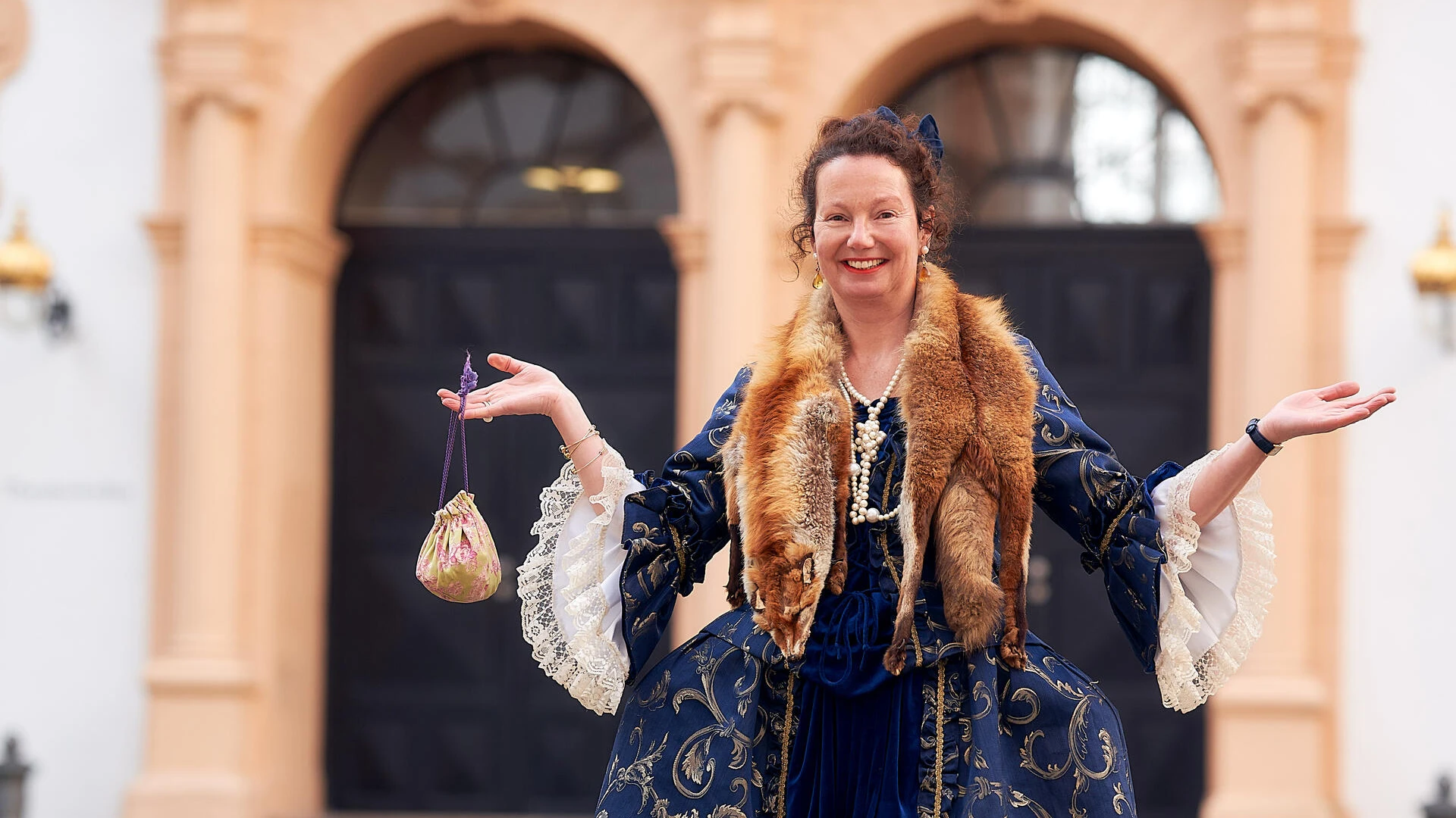 Gästeführerin als Lady Chitchat in historischem Kostüm vor dem Celler Schloss, lächeln und einladend gestikulierend.