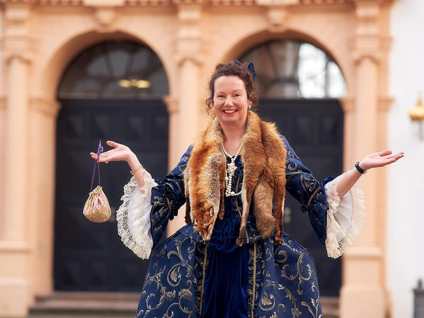 Gästeführerin als Lady Chitchat in historischem Kostüm vor dem Celler Schloss, lächeln und einladend gestikulierend.