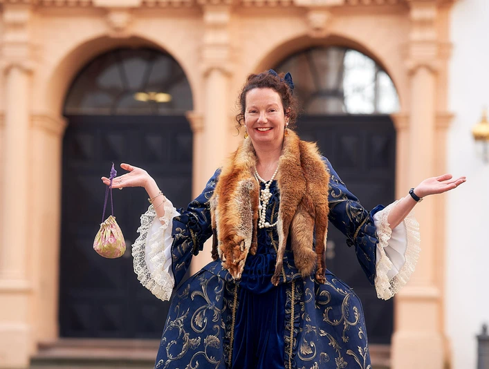 Lady Chitchat: Historische Kostümführung im Celler Schlos Gästeführerin als Lady Chitchat in historischem Kostüm vor dem Celler Schloss, lächeln und einladend gestikulierend.