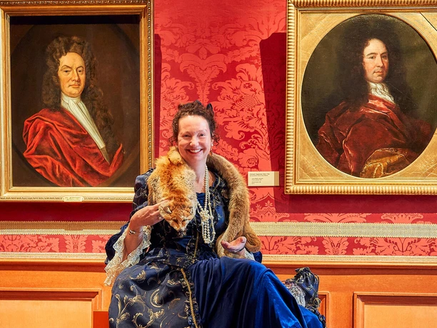Eine Frau in einem üppigen, blauen Renaissance-Kleid sitzt lächelnd vor zwei Porträts in einem prunkvollen Raum.