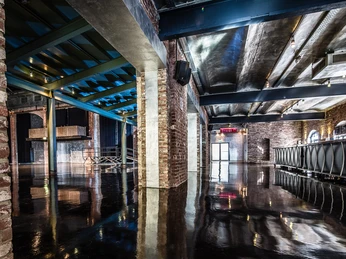 E-Werk Köln Foyer Das Bild zeigt das Foyer des E-Werks Köln mit industriellem Charme durch freiliegende Backsteinwände, einer glänzenden, polierten Bodenfläche und einer Decke mit sichtbaren Stahlträgern. Im Hintergrund sind moderne Lichtinstallationen zu erkennen.The picture shows the foyer of the E-Werk Cologne with its industrial charm thanks to exposed brick walls, a shiny, polished floor surface and a ceiling with visible steel beams. Modern lighting installations can be seen in the background.